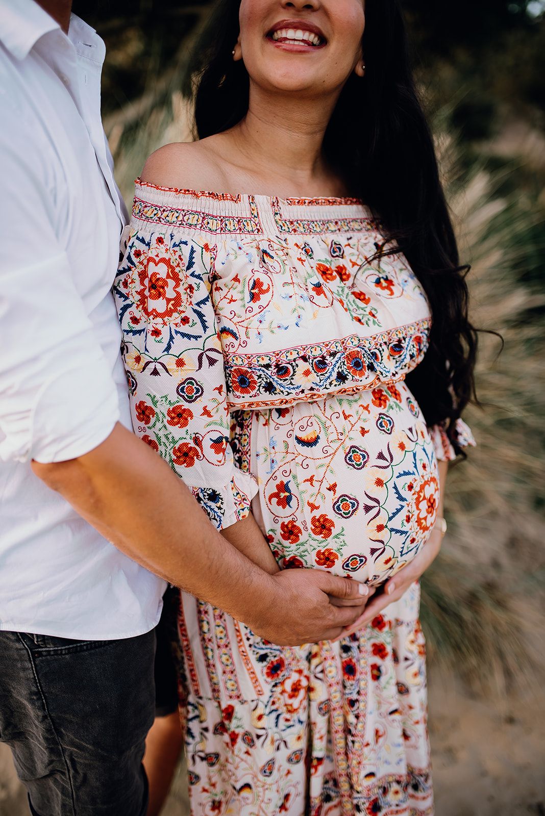 Pregnant woman in a floral off-shoulder boho dress cradling her baby bump outdoors with partner.