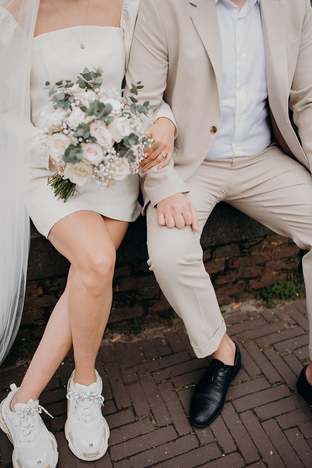 Bride holding white floral bouquet sitting beside groom in beige suit on brick steps.