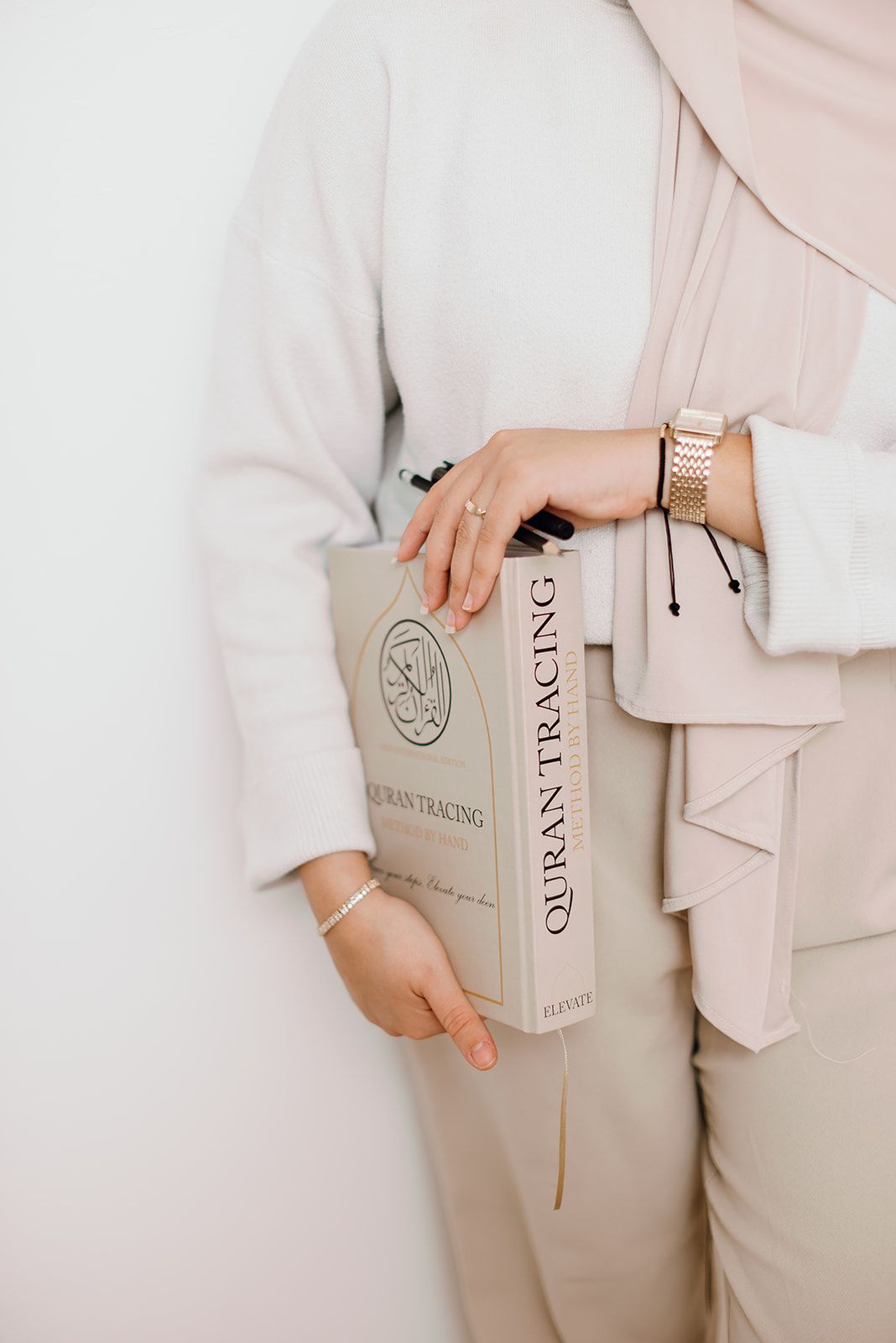 Woman in beige outfit holding a Quran Tracing book against a white wall.