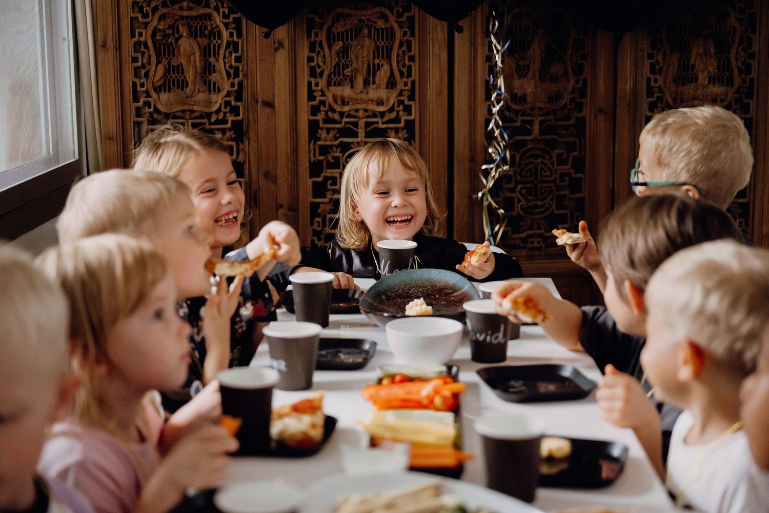 Happy children laughing and eating together at a festive birthday party table with colorful food.