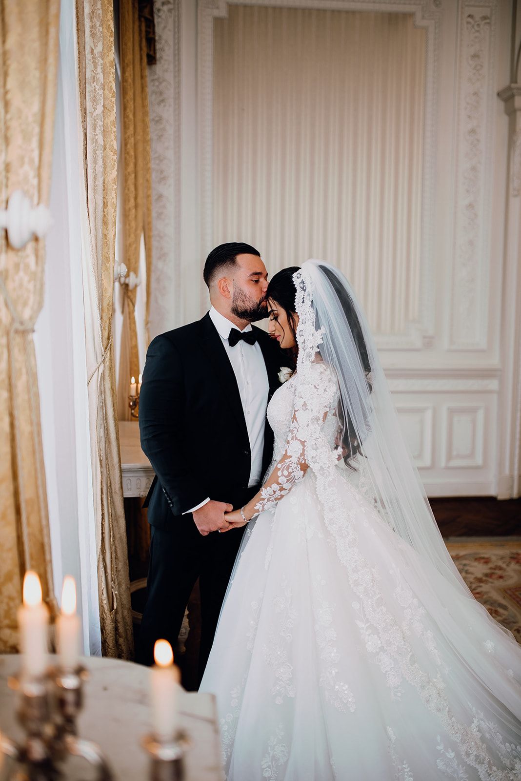 Groom in black tuxedo kisses bride in a lace ball gown inside an elegant candlelit room.