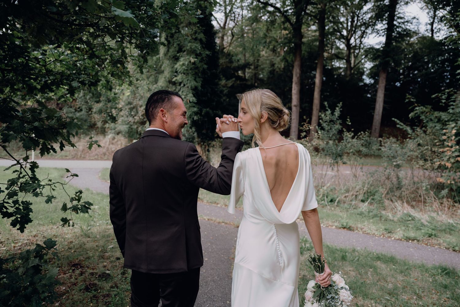 A groom in a dark suit kisses his bride's hand as she looks back in a white backless gown outdoors.