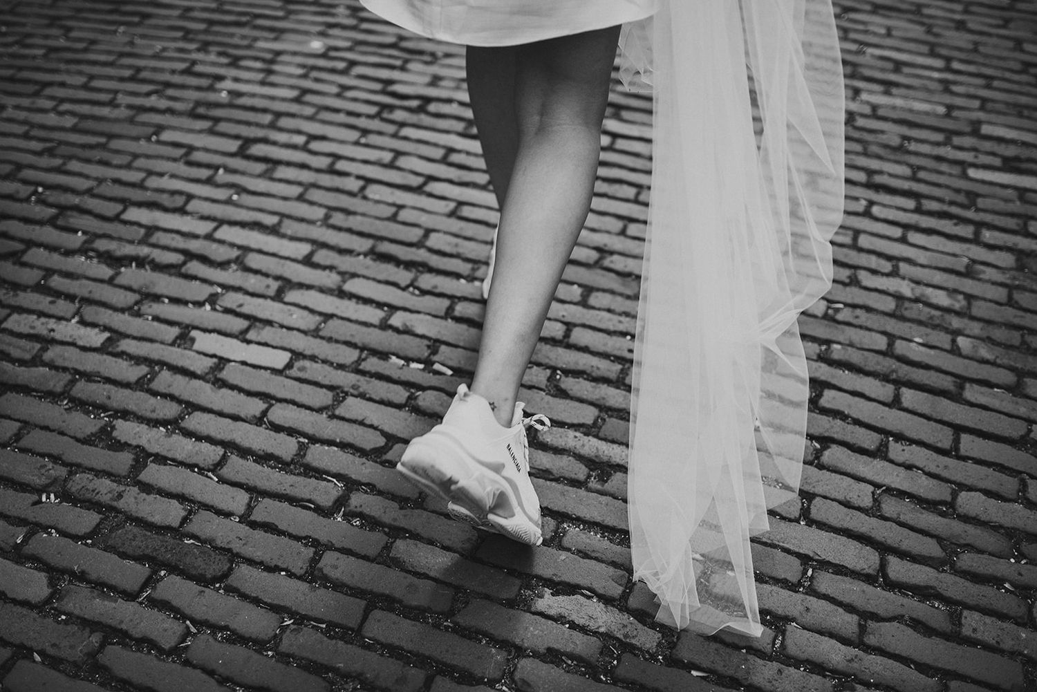 Black and white photo of a bride walking on cobblestones in white sneakers with wedding dress train.