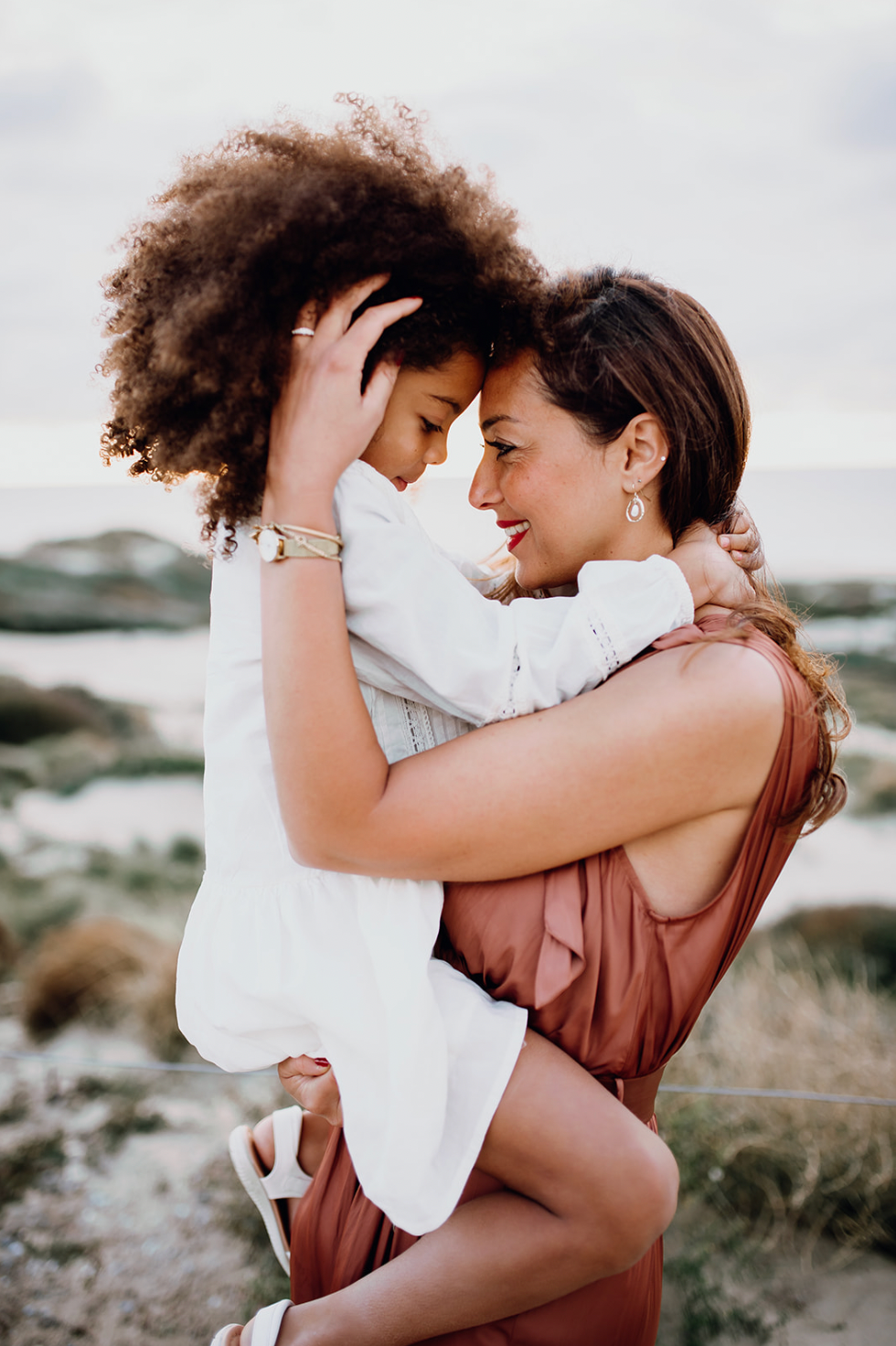 Mother smiling and holding young daughter close in a tender forehead-to-forehead embrace outdoors.