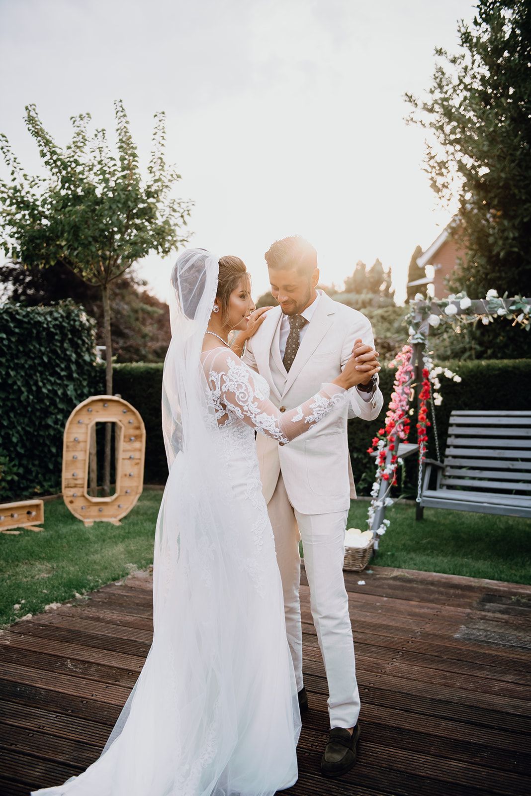 Bride and groom share first dance in sunlit garden, bride in lace veil gown, groom in white suit.