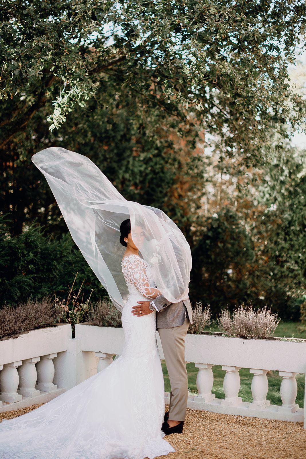 Bride in lace gown with flowing veil billowing in the wind beside groom in garden setting.