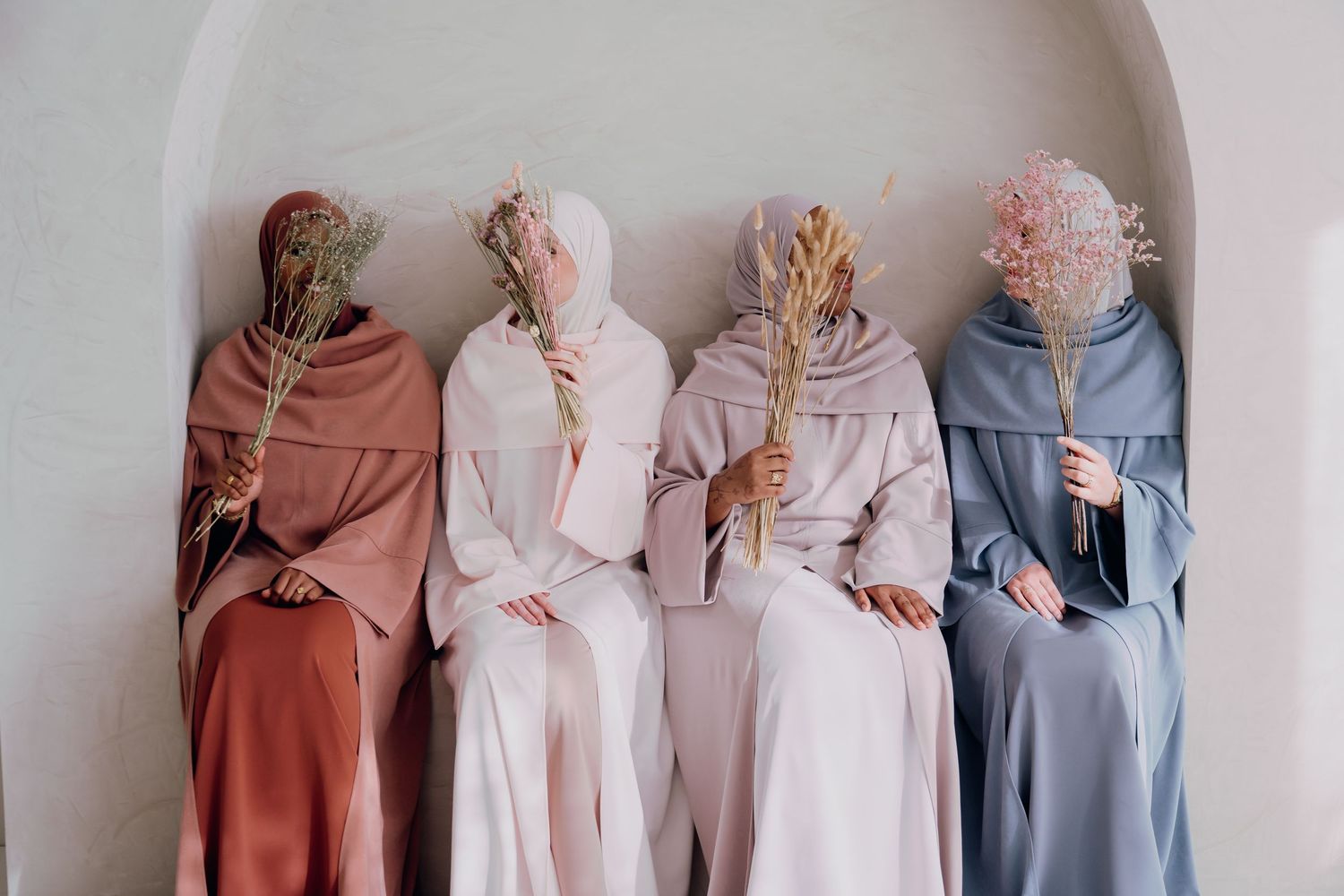 Four women in modest hijab attire hold dried flowers while seated in a neutral-toned architectural niche.