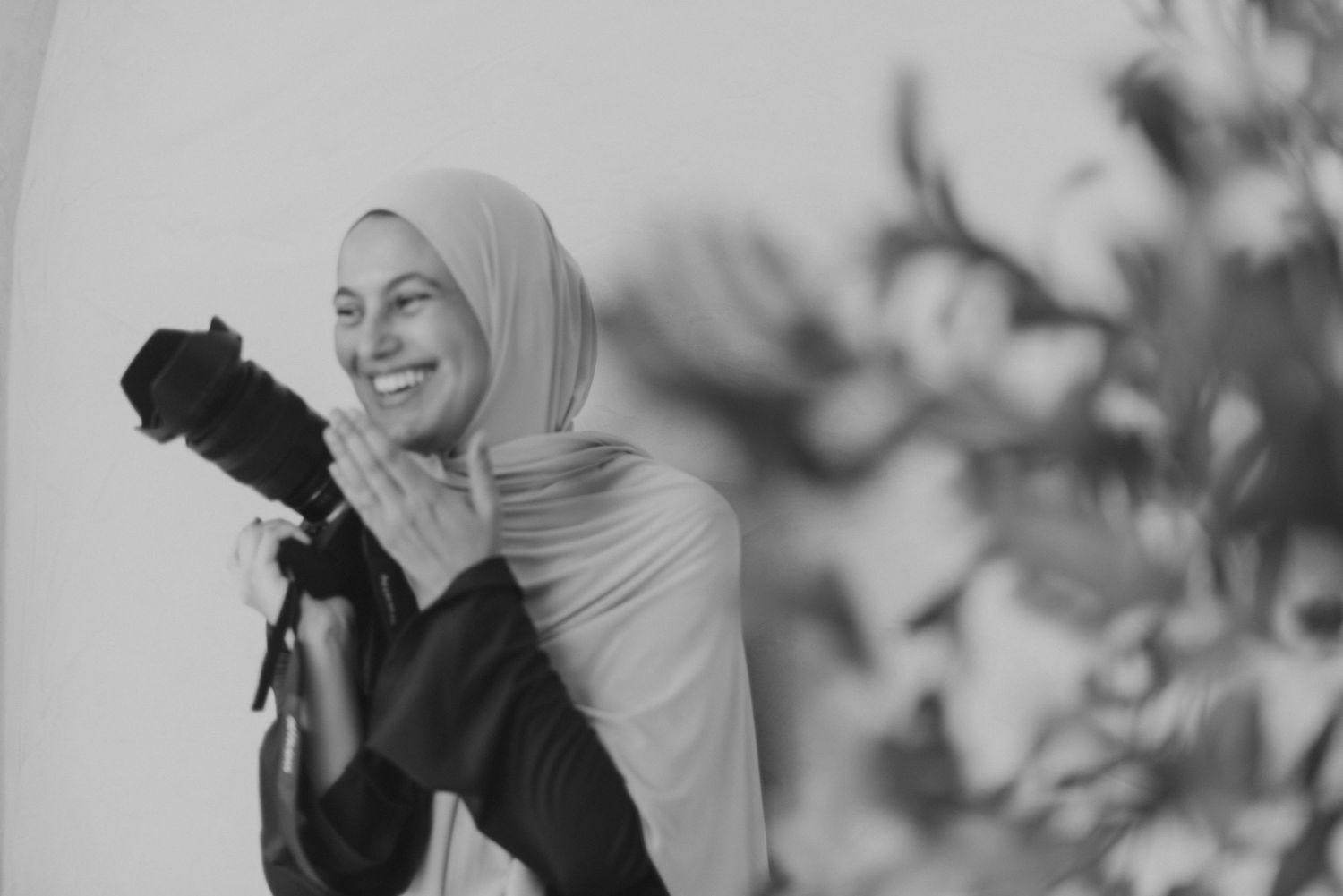 Smiling woman in hijab holding a professional camera, black and white photography portrait.