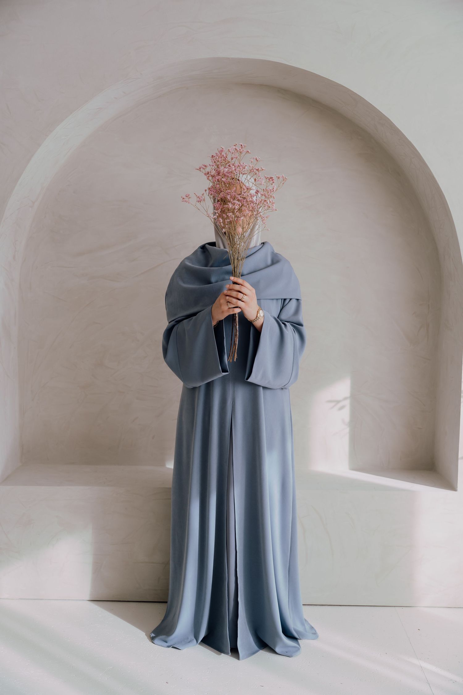 Person in flowing dusty blue abaya holding pink dried flowers in front of face, standing in minimalist white arched alcove.