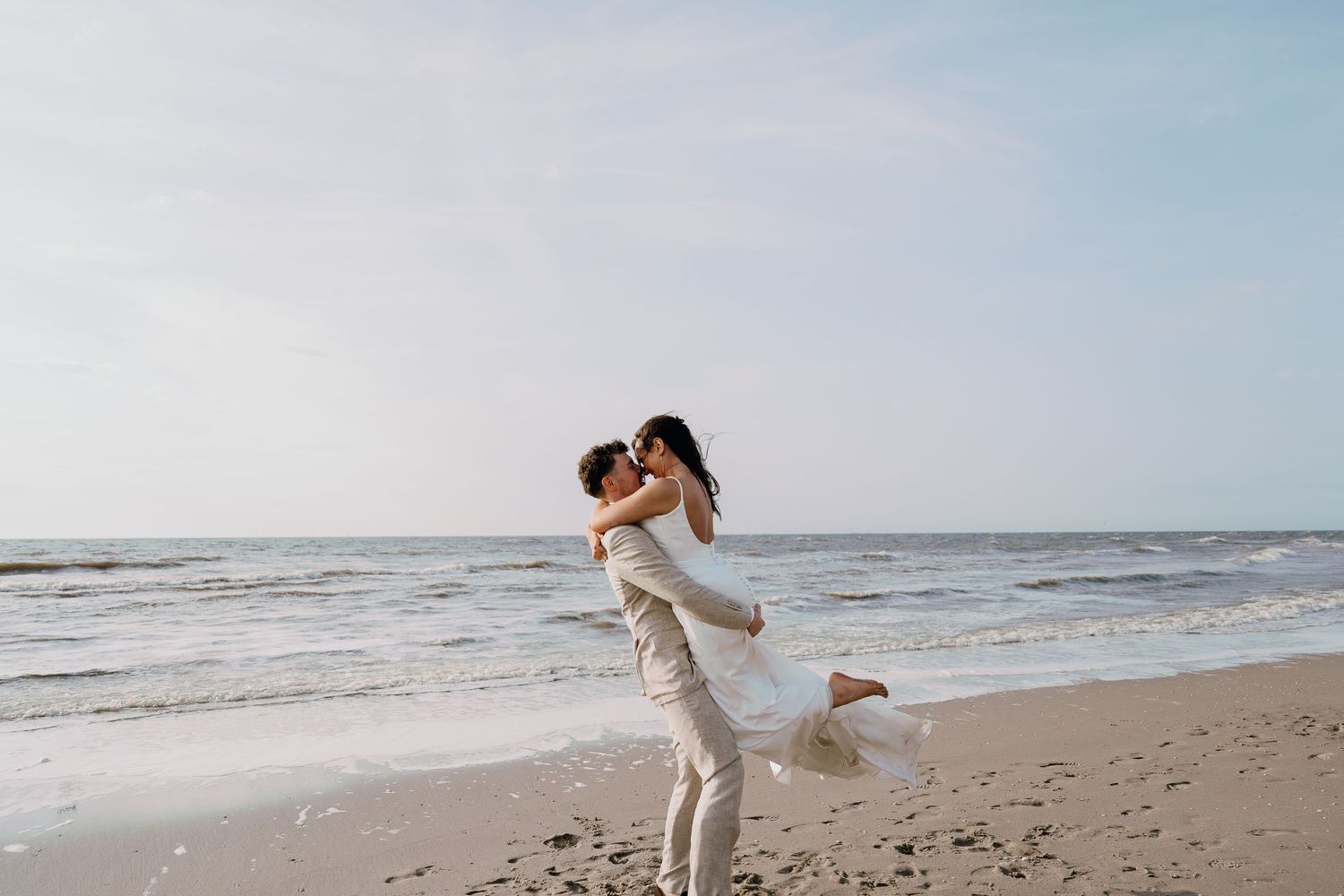 Couple sharing a romantic kiss on a sandy beach, groom lifting bride in wedding attire with ocean waves behind them.