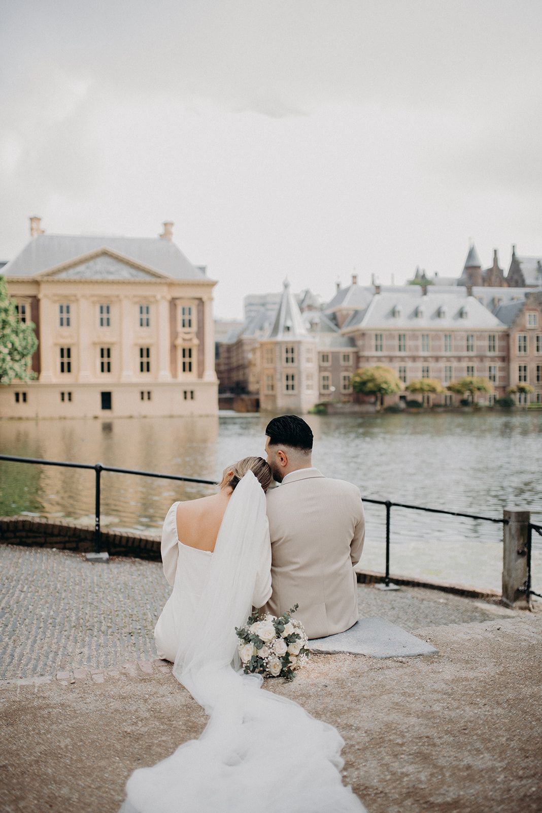 Bride and groom sitting by a waterfront with historic European buildings in the background.