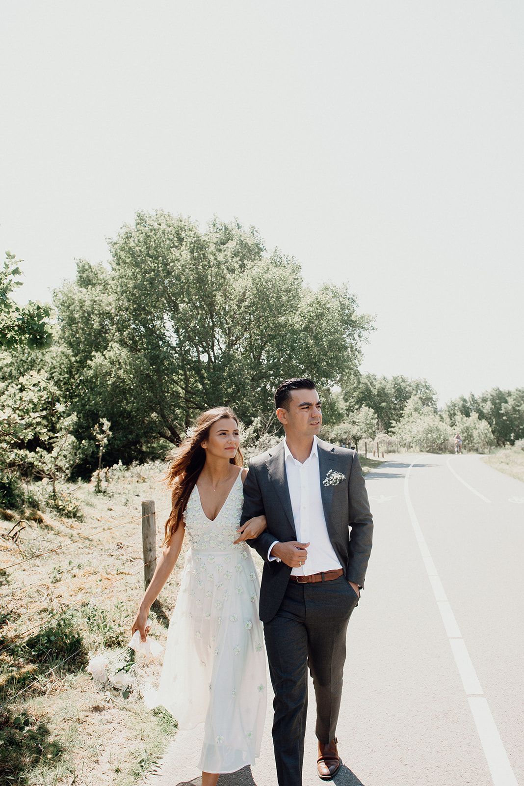 Bride in white lace dress and groom in dark suit walk arm in arm along sunny countryside road.