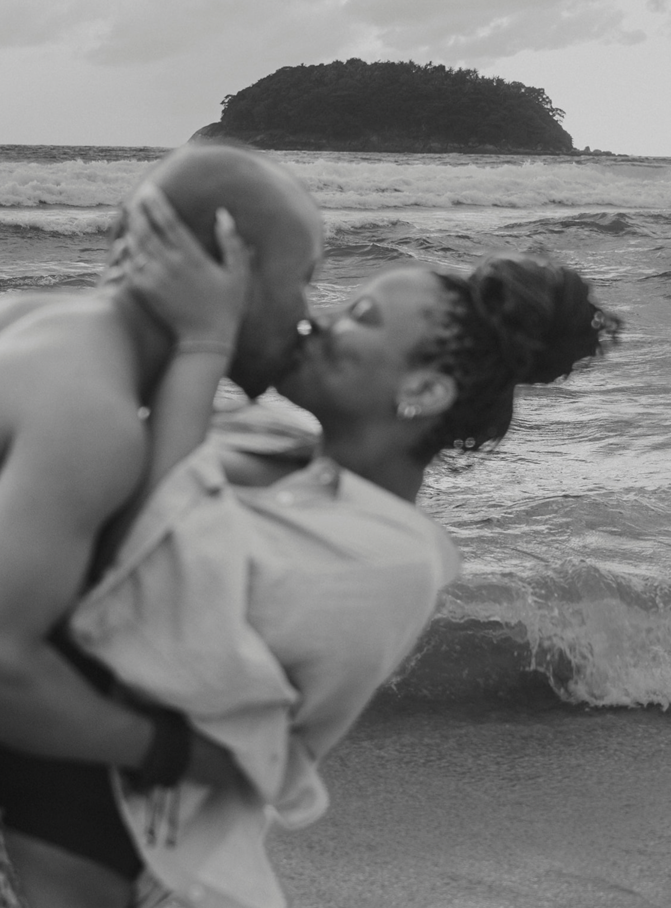 Black and white romantic beach kiss photo of a couple embracing by ocean waves with island in background.