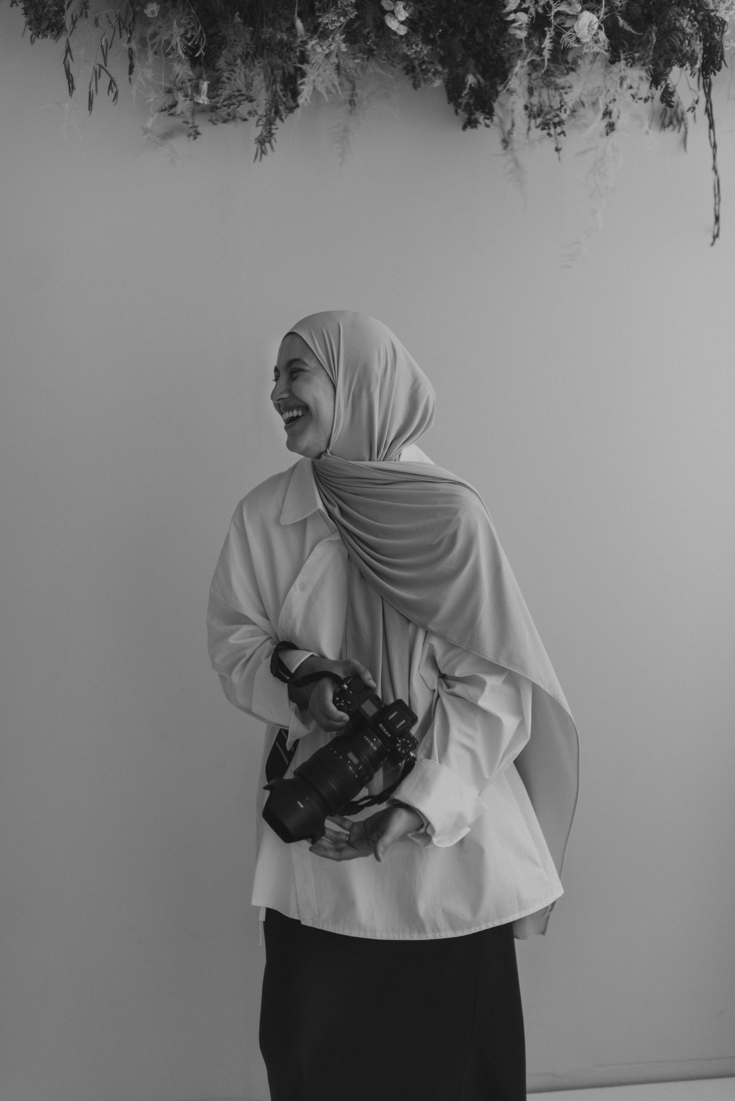 Black and white photo of a smiling woman in hijab holding a camera, standing beneath floral decorations.