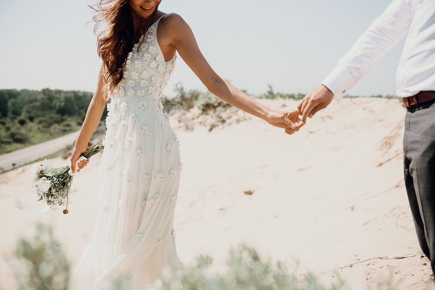 Bride in boho lace wedding dress holding bouquet walks hand in hand with groom across sandy dunes.