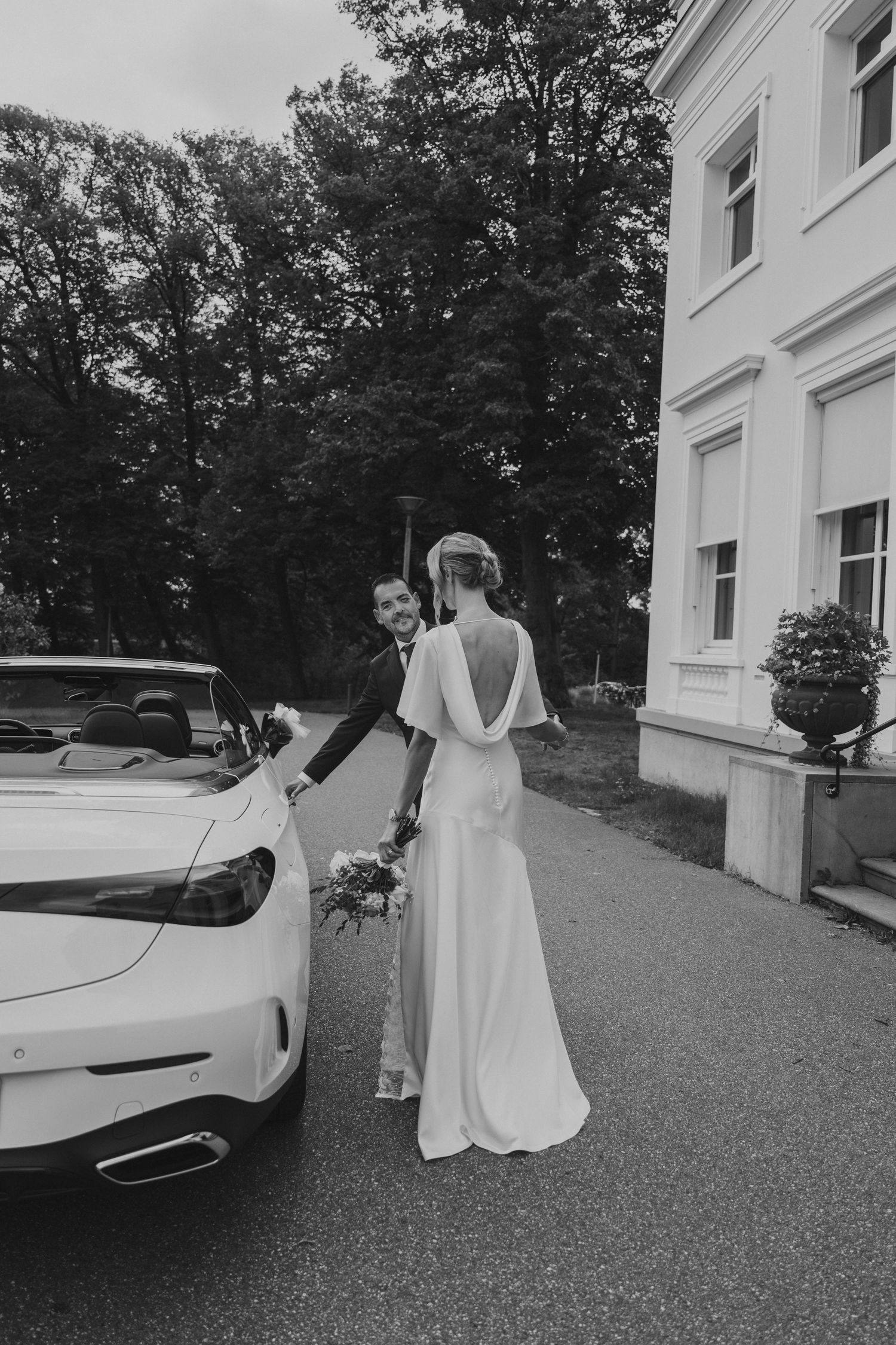 Black and white wedding photo of bride and groom beside a white convertible car outside an elegant estate.