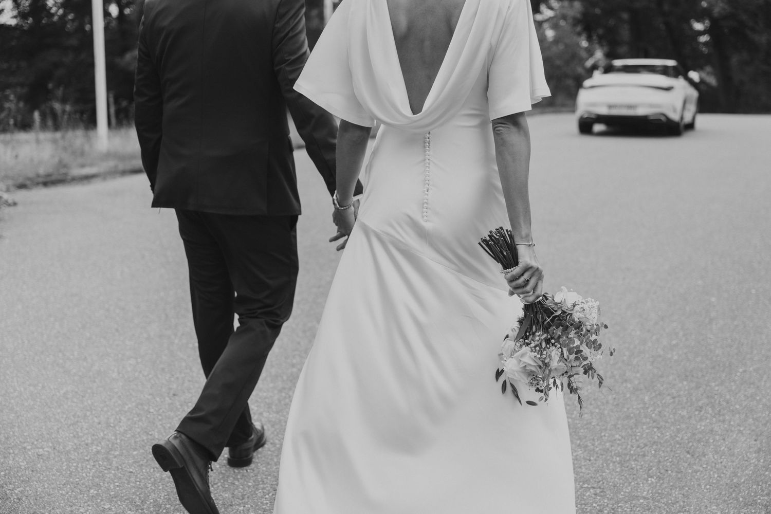 Black and white photo of a bride and groom walking hand in hand on a road, bride holding a bouquet.