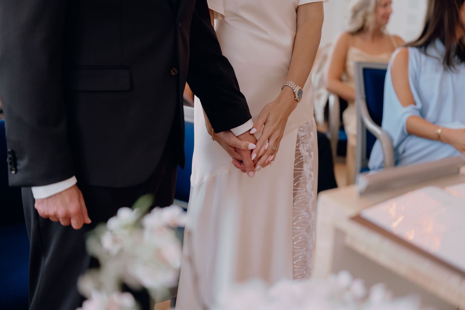 Bride and groom holding hands during wedding ceremony, bride in elegant white gown, guests seated in background.
