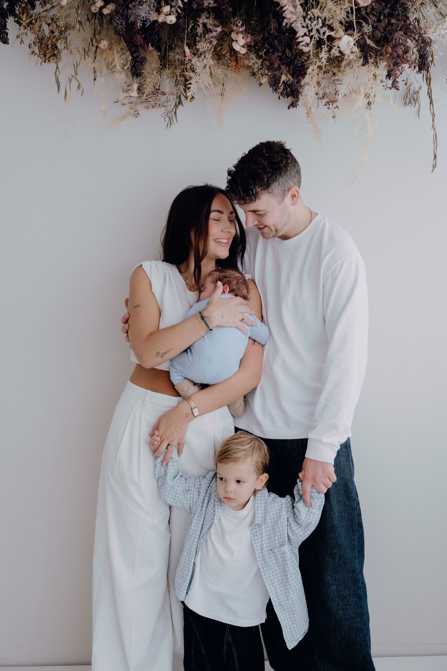 A young family poses tenderly with a newborn baby and toddler under dried floral decor in a bright studio.