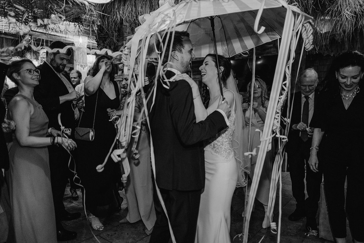 Black and white photo of bride and groom kissing under decorated umbrella surrounded by cheering guests. Jufenalm