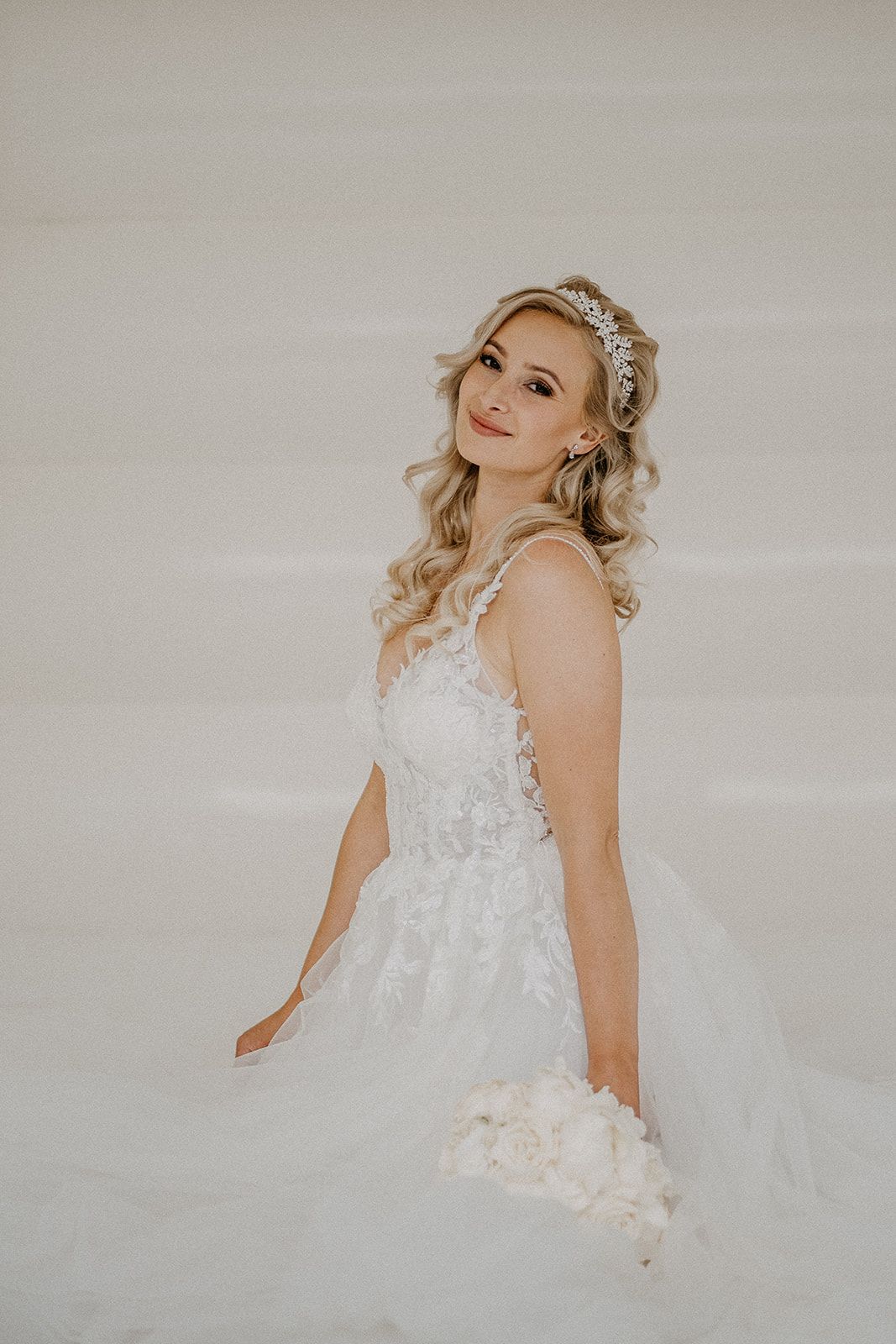 Beautiful blonde bride in a lace wedding gown and tiara smiling softly in a white bouncy castle.