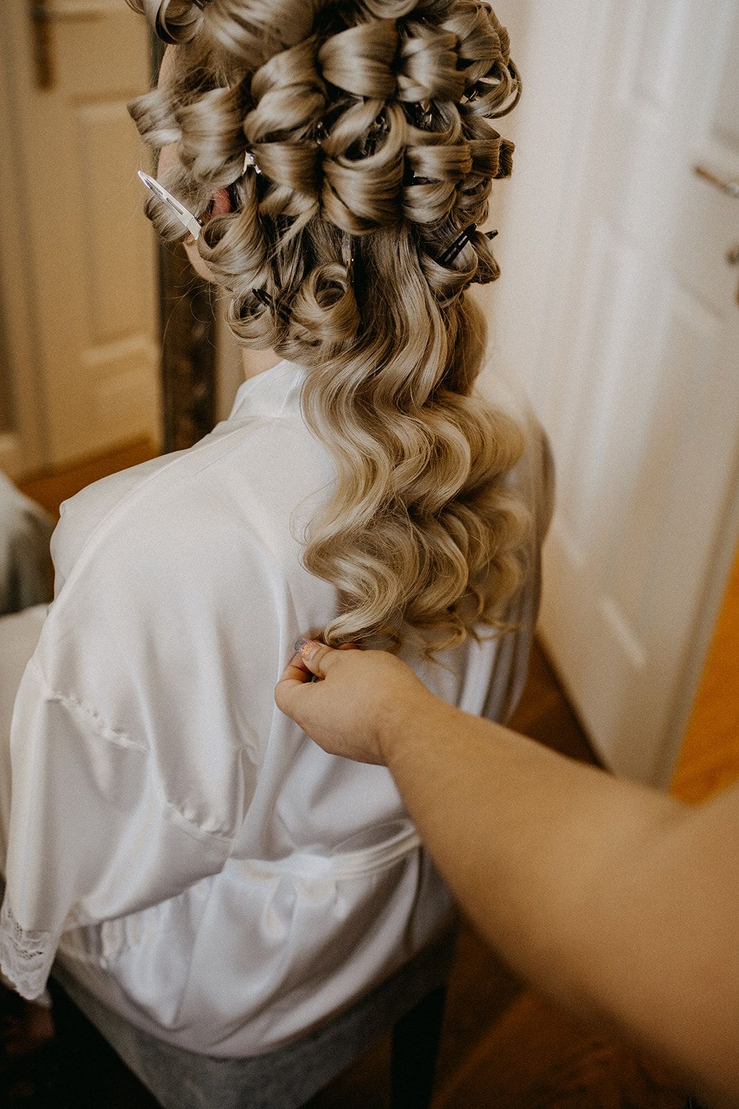 Bridesmaid fastens the back of a brides elegant curled updo hairstyle in a white satin robe.