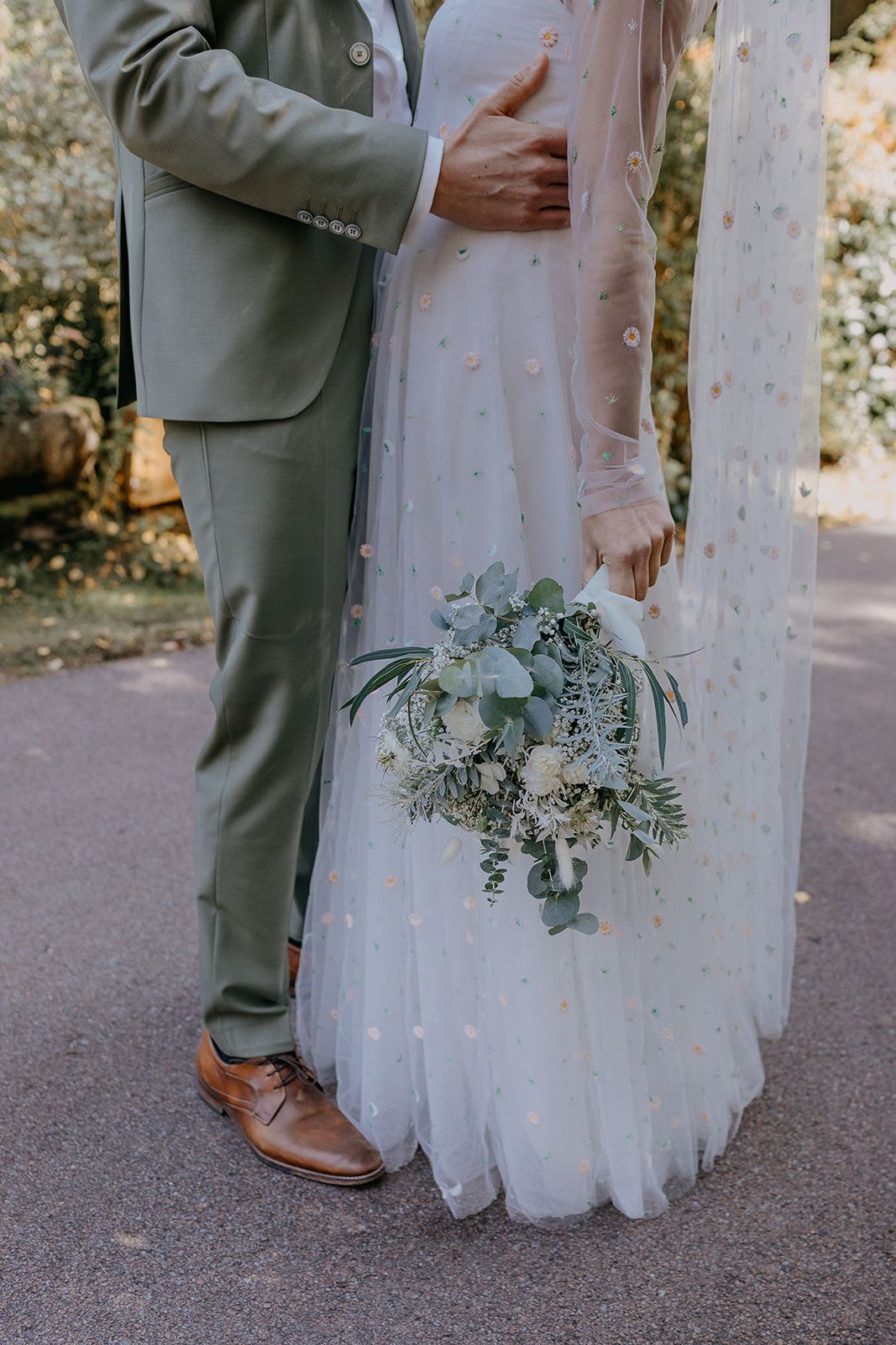 Bride and groom embracing outdoors, bride holding eucalyptus bouquet, groom in sage green suit.