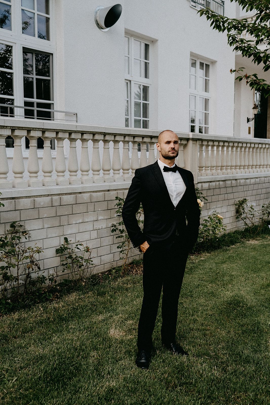 Dapper groom in black tuxedo and bow tie stands confidently on lawn outside a white building.