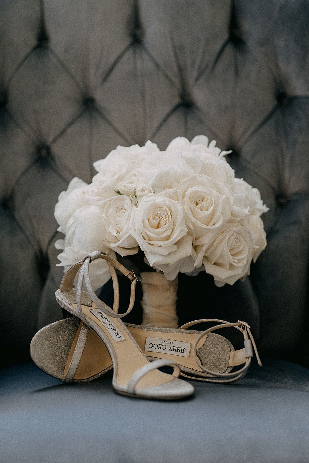 Jimmy Choo gold glitter bridal heels displayed with a lush white rose bouquet on tufted gray chair.
