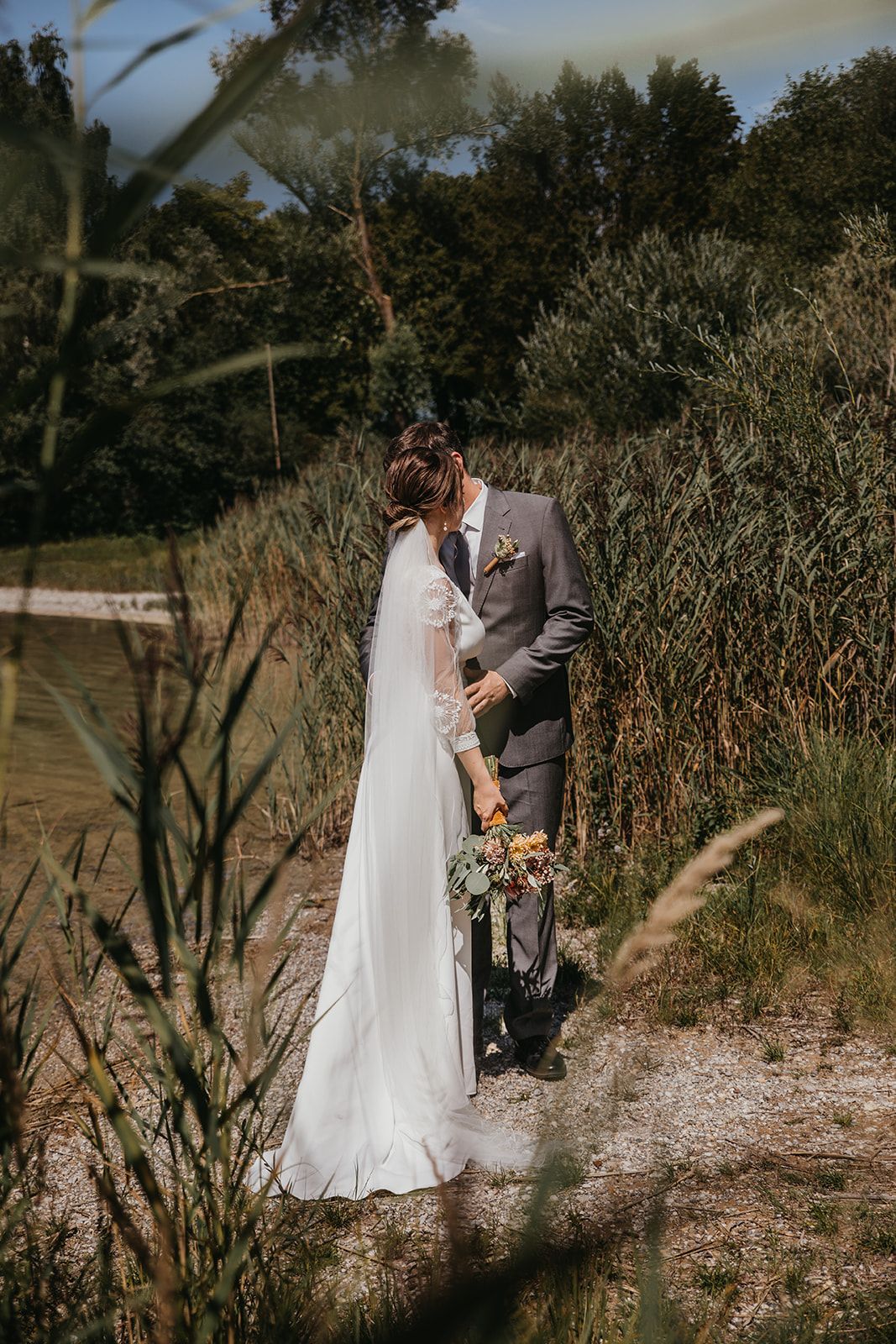 Bride and groom sharing a kiss near a river surrounded by tall reeds and greenery.