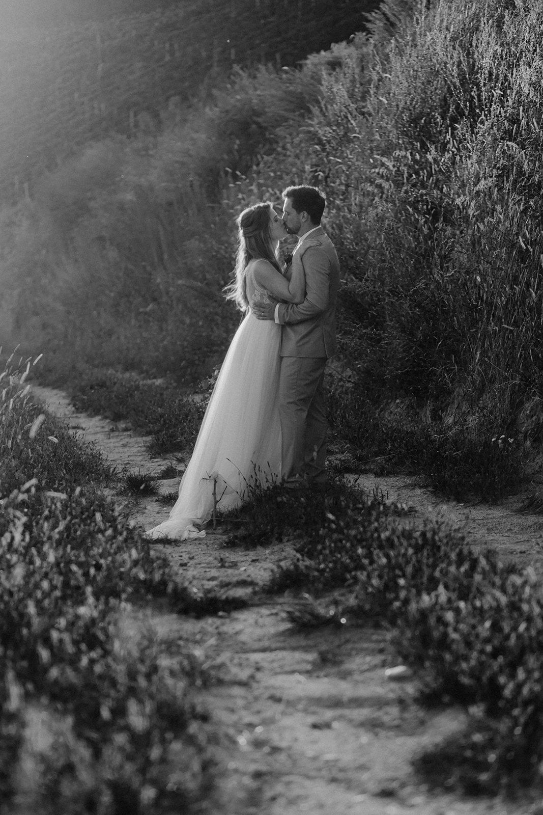 Black and white photo of bride and groom kissing on a rural path surrounded by wildflowers.