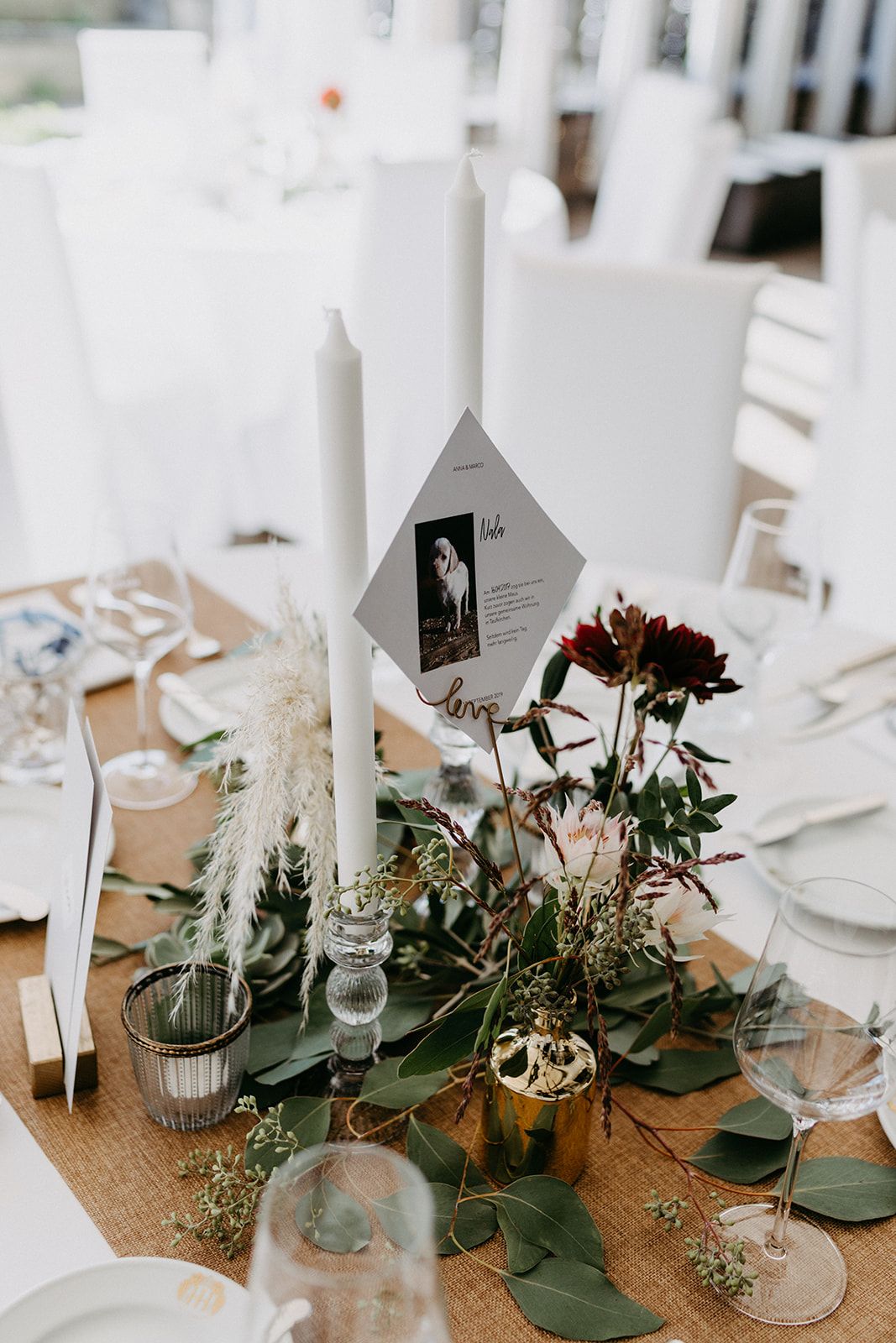 Elegant wedding table centerpiece with white taper candles, eucalyptus greenery, burgundy flowers, and a table number card.