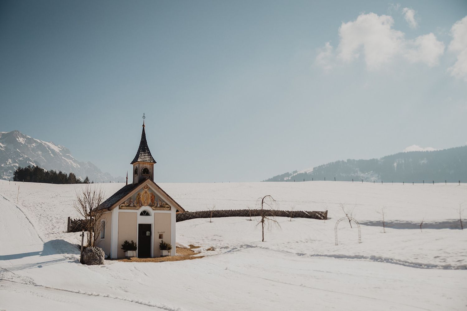 A small white chapel surrounded by a vast snowy winter landscape with mountains in the background. Jufenalm