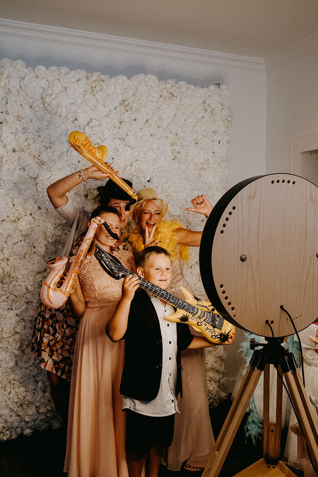 Wedding guests pose with inflatable instruments in front of a white flower wall at a photo booth.