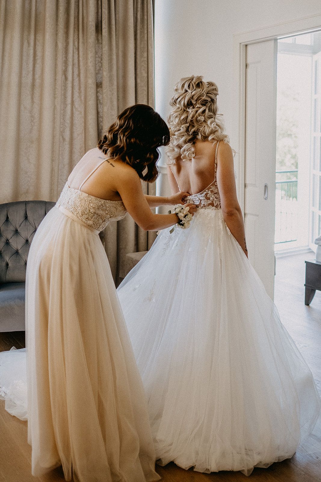 Bridesmaid in lace gown helps fasten the back of a brides flowing white ball gown wedding dress.