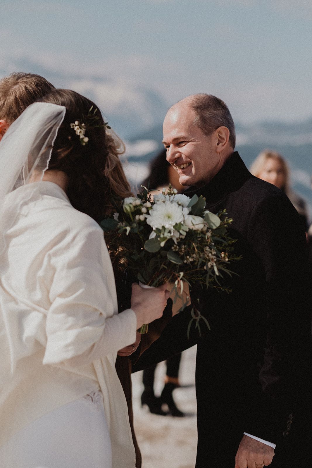 Smiling groom in black suit holds white bouquet facing bride in white dress and veil at snowy mountain wedding ceremony. Jufenalm