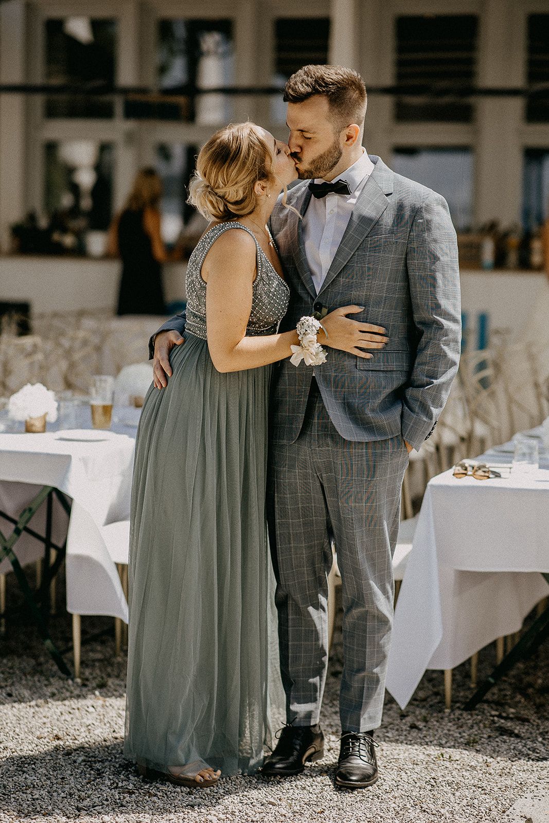 Couple sharing a kiss at wedding reception, woman in sage green gown with wrist corsage, man in grey plaid suit with bow tie.