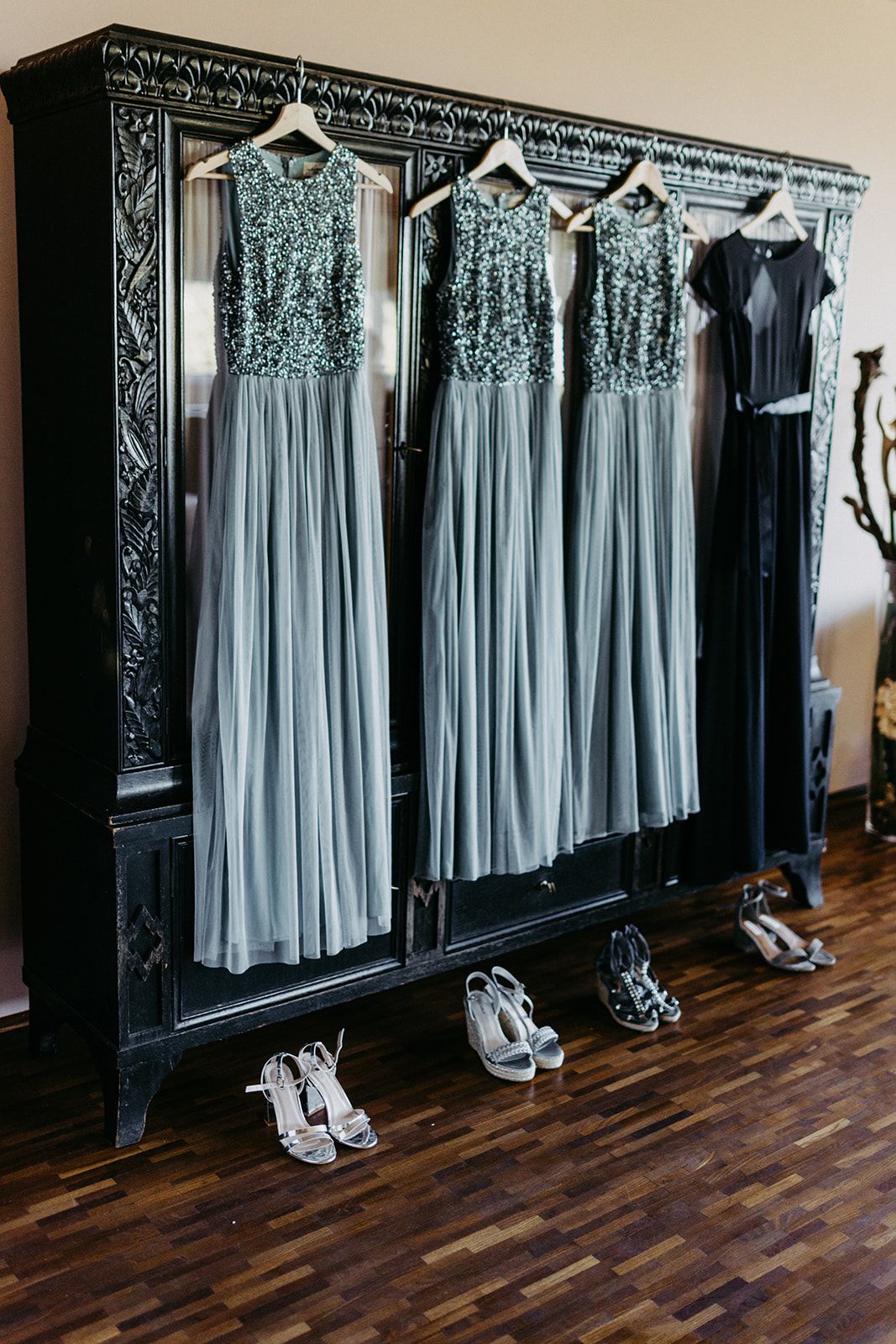 Three sage green sequin bridesmaid dresses hanging on ornate black wardrobe with matching shoes below.