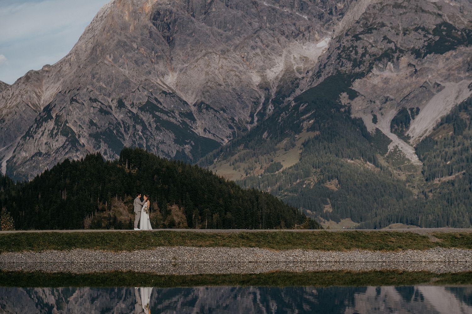 Wedding couple kissing by a reflective alpine lake with dramatic mountain backdrop.