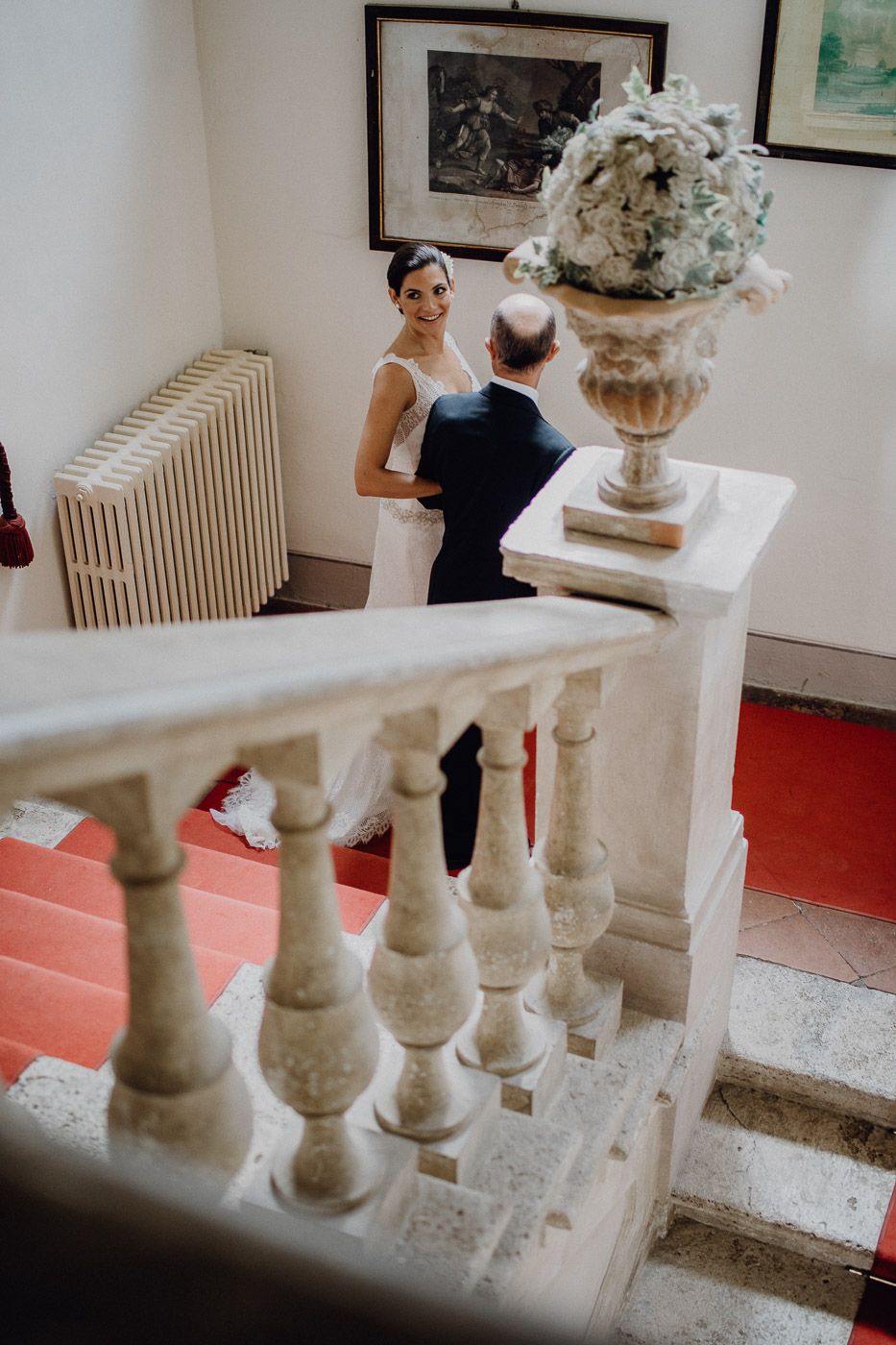 Bride in white dress smiles at older man near ornate marble staircase with floral arrangement. Tuscany Villa Catignano