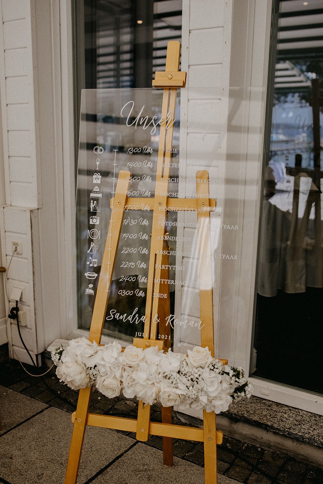Elegant acrylic wedding timeline sign on wooden easel decorated with white roses.