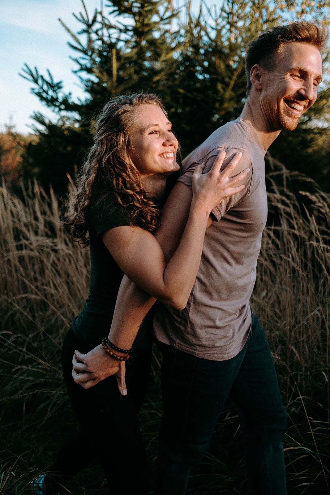 Happy couple laughing and embracing in golden hour sunlight among tall grass and evergreen trees.