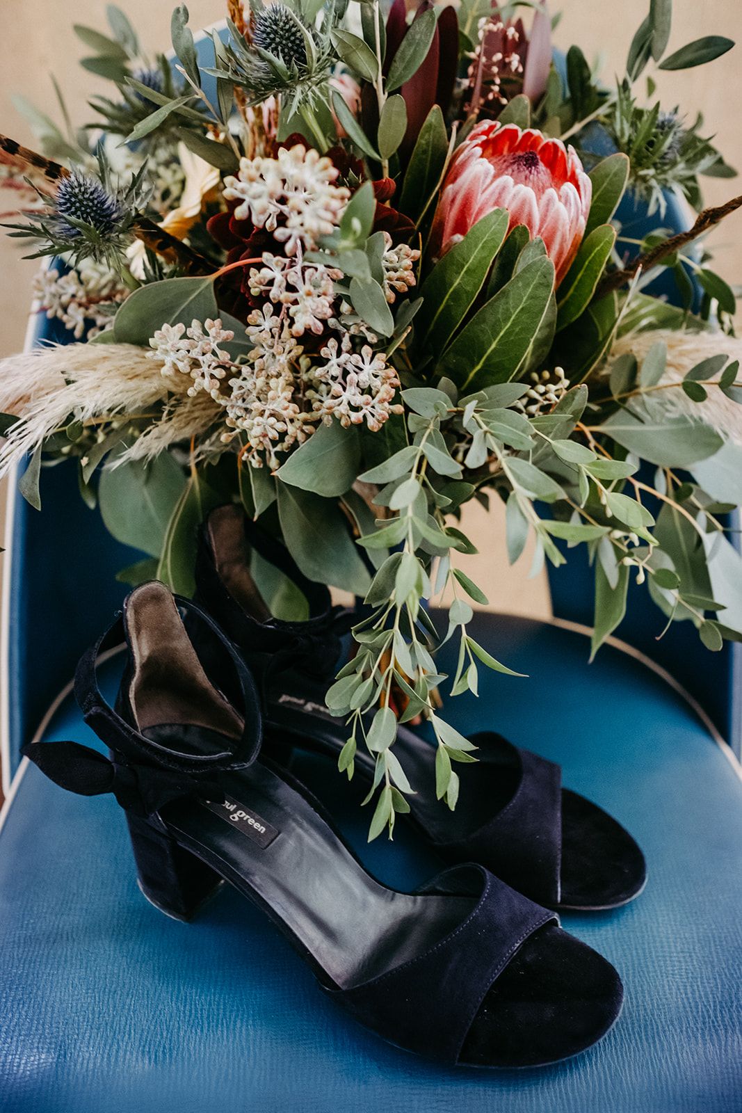 Navy suede block heel sandals beside a wild boho bridal bouquet with protea on blue chair.