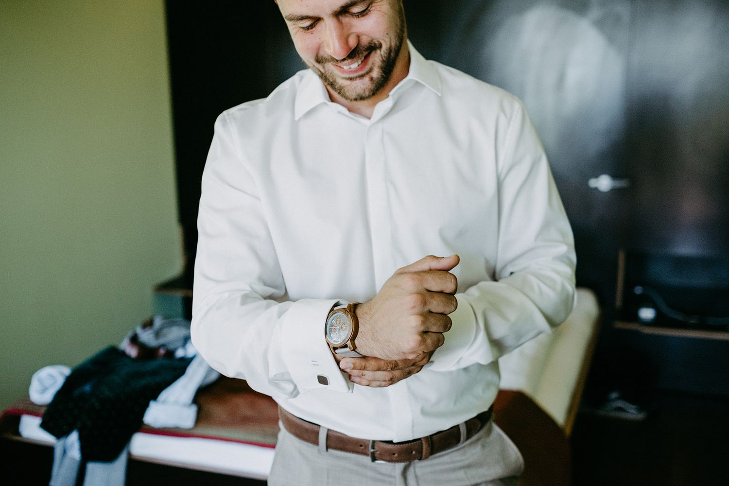 Smiling groom in white dress shirt adjusting his watch while getting ready for his wedding.