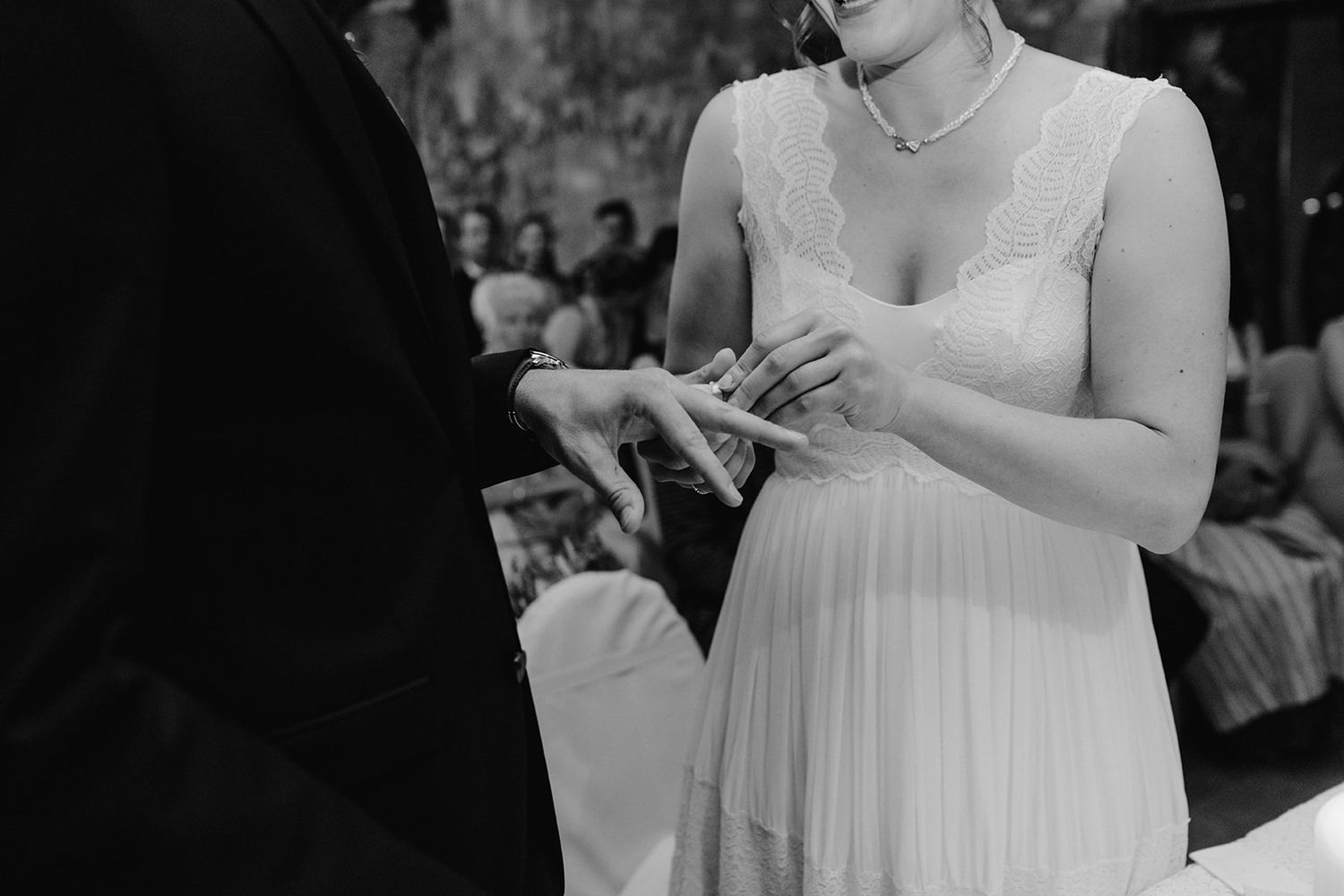 Black and white close-up of a bride in a lace wedding dress placing a ring on the grooms finger.