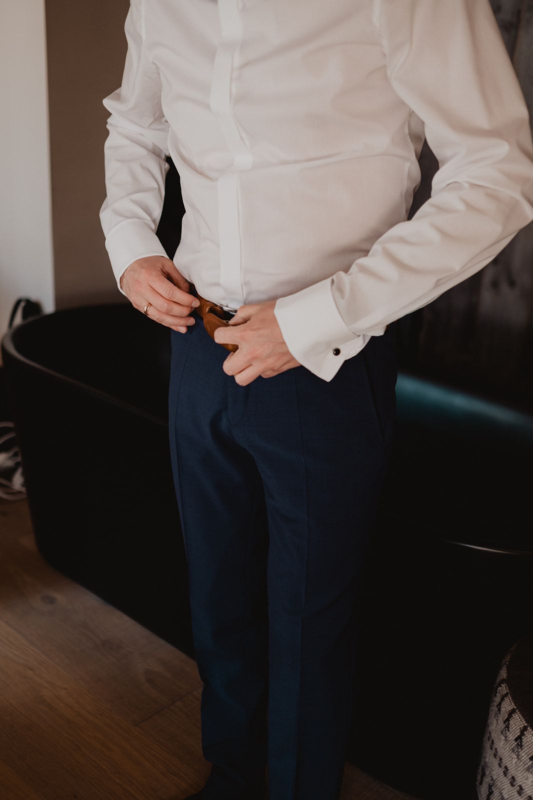 Groom in white dress shirt and navy trousers fastening his belt while getting ready for wedding.