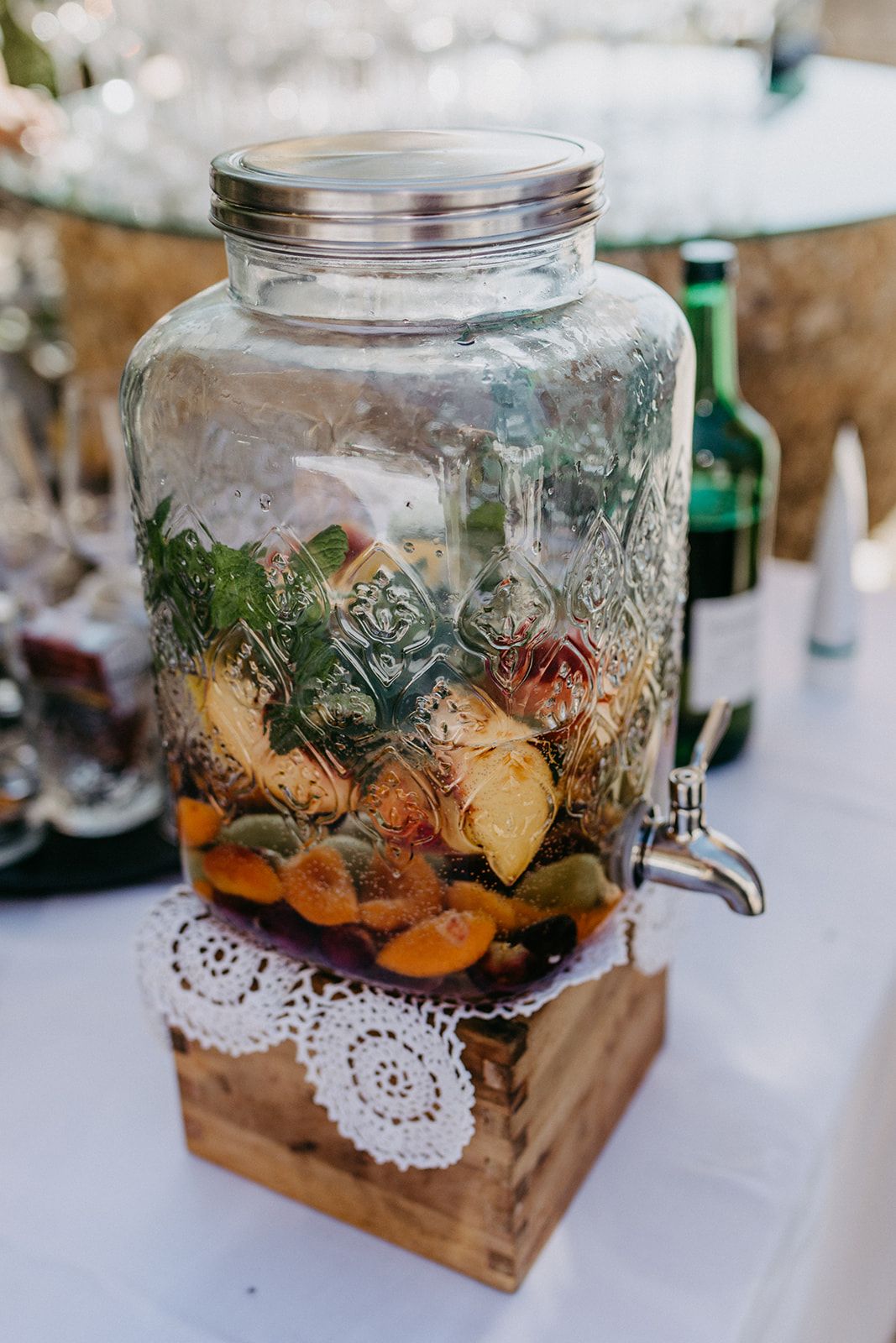Decorative glass beverage dispenser filled with colorful fruits on a wooden stand at wedding reception.