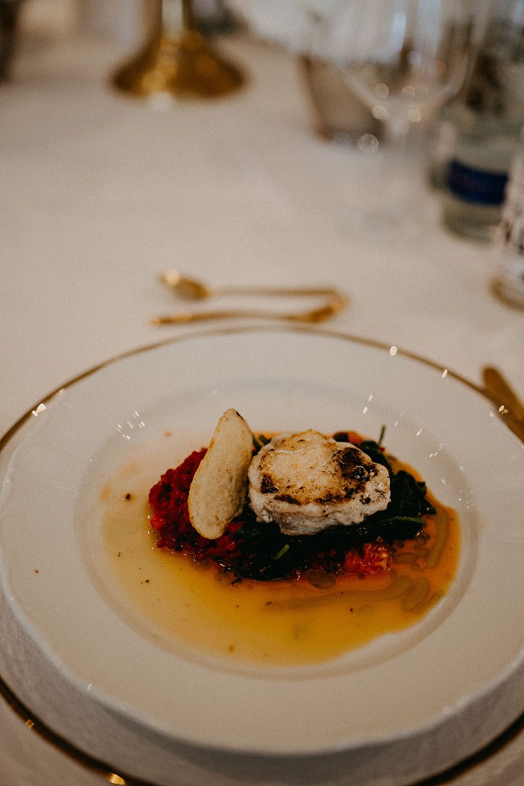 Elegant plated starter with goat cheese, dark greens, tomato sauce, and bread on a white gold-rimmed plate.