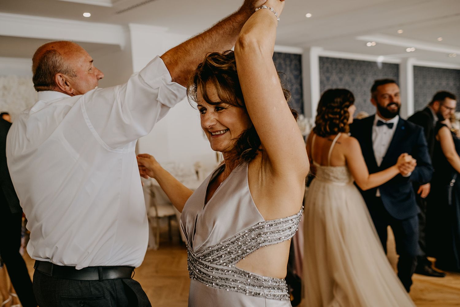 Woman in silver beaded dress dancing joyfully with arms raised at a wedding reception.