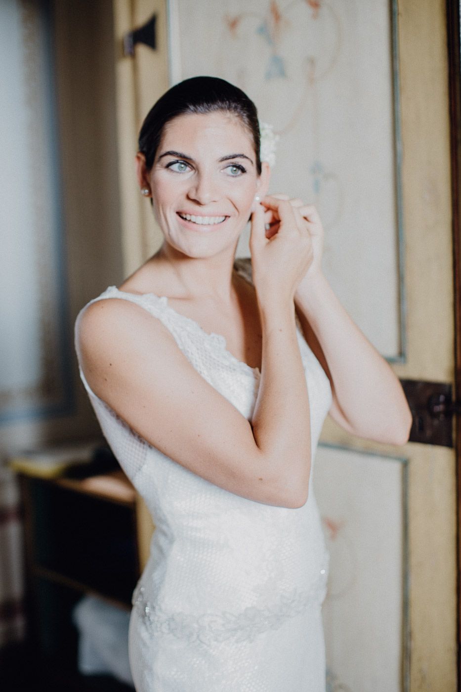 Beautiful smiling bride in white lace gown puts on earrings during wedding day preparations. Tuscany Villa Catignano